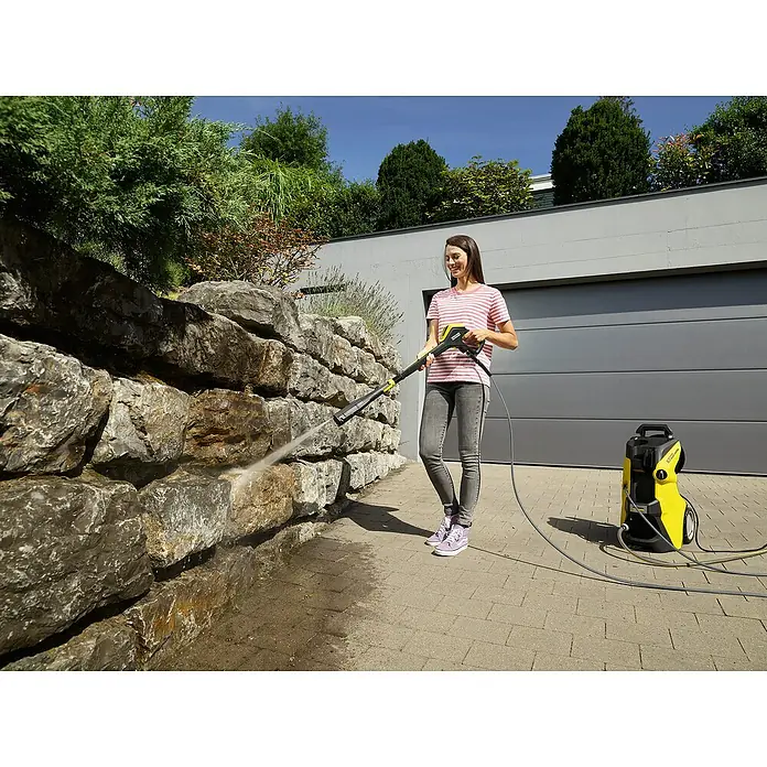 Kärcher Hidrolimpiadora de alta presión K7 Premium Smart Control Flex - 180 bar, 3.000 W, Capacidad de bombeo: 600 l/h, 5 pzs. - Mujer limpiando pared de piedra con limpiadora de alta presión Kärcher amarilla, que está de pie junto a ella. Ideal para la limpieza de casa.
