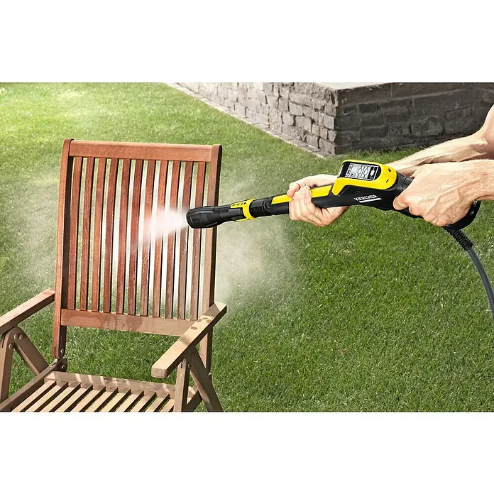 Kärcher Hidrolimpiadora de alta presión K 7 Smart Control Flex - 180 bar, 3.000 W, Capacidad de bombeo: 600 l/h, Sin Home Kit - Un hombre limpia una silla de madera al aire libre con una hidrolimpiadora amarilla Kärcher, el chorro de agua elimina la suciedad.