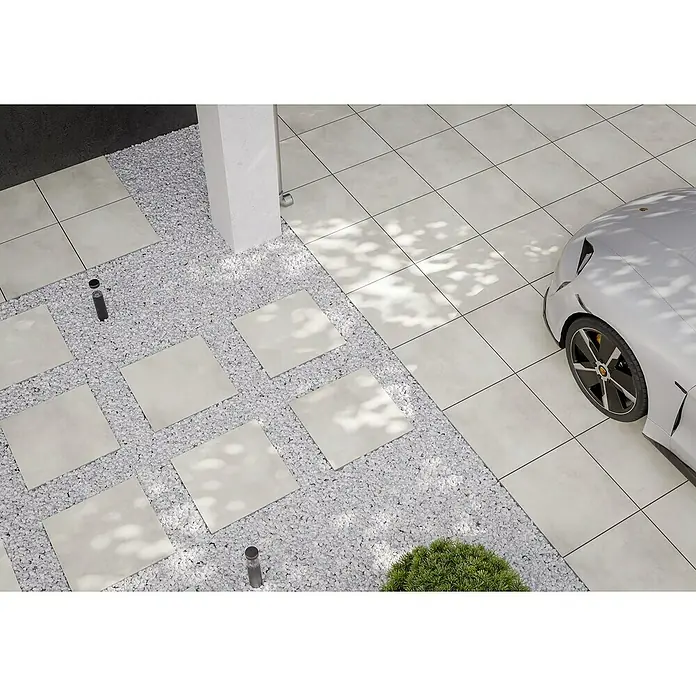 Terrassenfliese Dimension - 59,8 x 59,8 x 2 cm, Beige, Matt - Ein Außenbereich mit hellen Fliesen und einem Beet mit weißen Steinen, durch das ein Plattenweg führt. Ein stilvoller Carport mit Auto ist erkennbar.