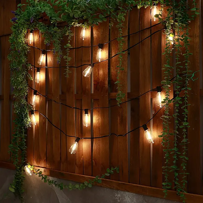 Arte confort Lámpara decorativa solar Glimia - Para exterior, 785 cm, Blanco cálido - Luces cálidas realzan una pared de madera con enredaderas verdes, creando un ambiente acogedor.