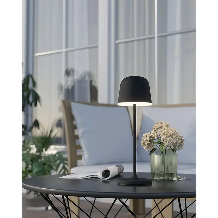 Eglo Lámpara de sobremesa a batería Mannera - Ø x Al: 11 x 34 cm, Negro, 2,2 W, IP54 - Elegante lámpara de mesa negra que ilumina una mesa redonda, junto a un jarrón con flores y un periódico, ofreciendo iluminación elegante.