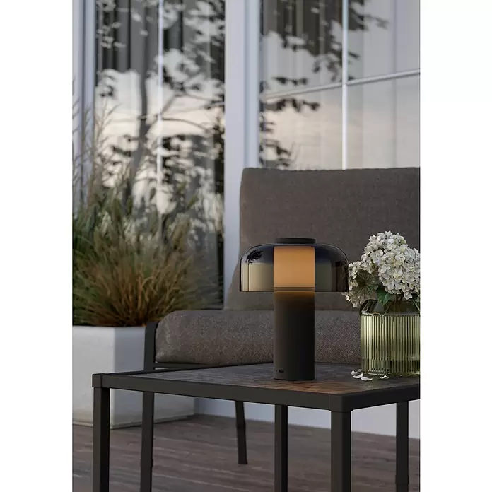 Lámpara de sobremesa LED redonda Ponente - 2,25 W, Ø x Al: 16 x 20 cm, Negro, Blanco cálido - Lámpara de mesa portátil negra con luz ámbar, en una mesa de patio exterior junto a una silla y flores.