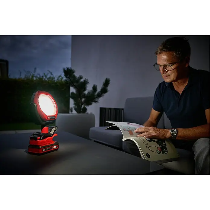 Einhell Power X-Change 18V Akku-Lampe GC-OL 18/1500LI SOLO - 18 V, Li-Ionen, Ohne Akku - Einhell Akku-Arbeitsleuchte erhellt eine Terrasse im Dunkeln, während ein Mann ein Buch liest. Die Lampe ist rot und schwarz und scheint hell.