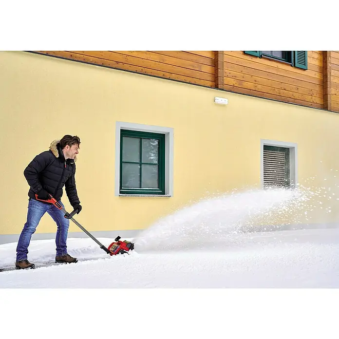Einhell Power X-Change 18V Akku-Schneefräse GE-ST 36/40LI E - 36 V, Ohne Akku, Arbeitsbreite: 40 cm - Ein Mann benutzt eine rote Schneefräse, um Schnee vor einem gelben Gebäude mit Fenstern zu räumen.