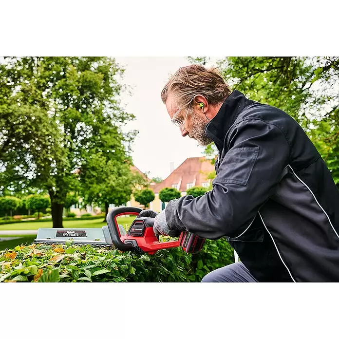 Einhell Professional PXC 18V Cortasetos de batería SOLO GP-CH 18/50 LI BL - 18 V, Iones de litio, Sin batería, Longitud de corte: 50 cm - Un hombre recorta un seto verde con un cortasetos Einhell rojo y negro, usando gafas de seguridad y tapones para los oídos.