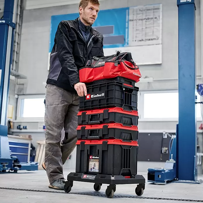Einhell Transportroller E-Case - L x B x H: 53 x 40 x 11 cm, Polypropylen, Traglast: 120 kg - Mann schiebt einen Stapel schwarzer und roter Einhell E-Case System Werkzeugkoffer auf einem Rollwagen in einer Werkstatt.
