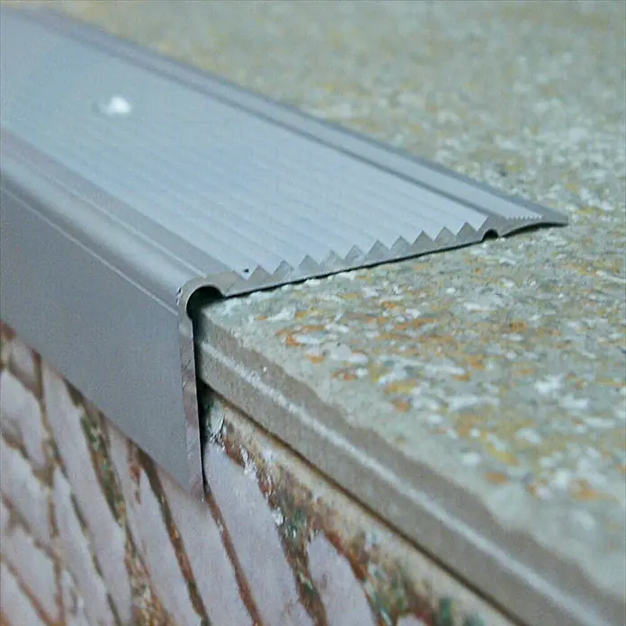Escalón autoadhesivo - 1 m x 45 mm x 22 mm, Aluminio - Primer plano de un perfil de borde de escalera plateado sobre escalones de baldosas, diseñado con una superficie ranurada para un agarre seguro.