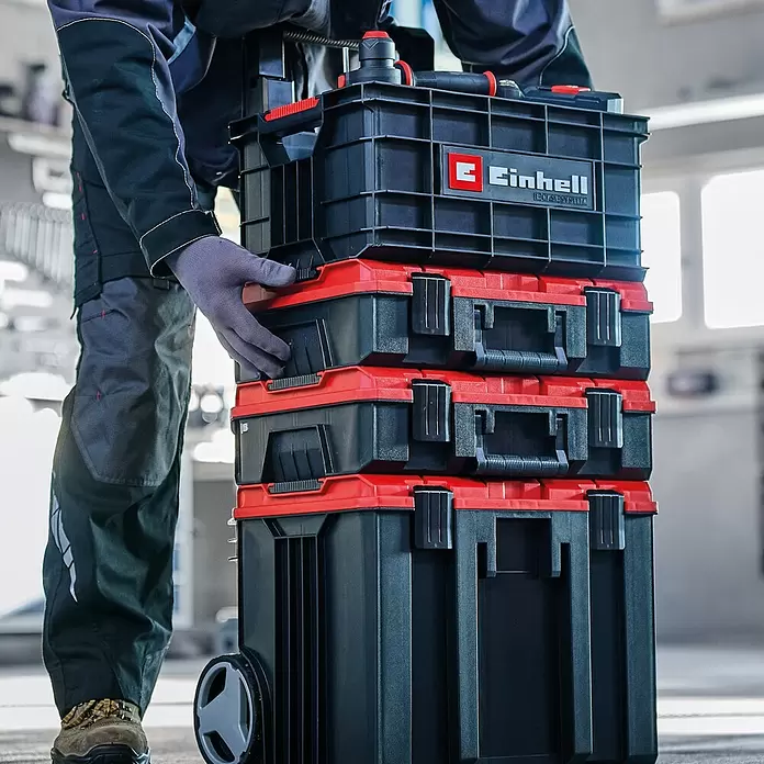 Einhell Werkzeugkasten - L x B x H: 32 x 44,4 x 21 cm, Polypropylen, Traglast: 90 kg - Person handhabt einen Stapel schwarzer und roter modularer Werkzeugkoffer auf Rollen. Das Einhell System ist tragbar und stapelbar.