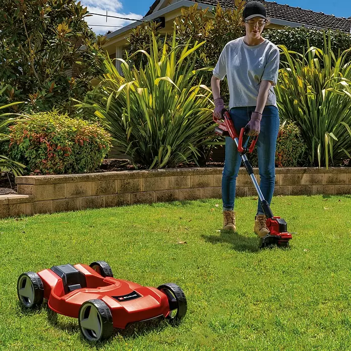 Einhell PXC 18V Akku-Rasentrimmer GE-CT18/28LI TC-Solo - 18 V, Li-Ionen, Ohne Akku, Schnittbreite Faden: 28 cm - Eine Person trimmt Gras mit einem roten Akku-Rasentrimmer; ein roter Mähroboter steht daneben auf dem Rasen.