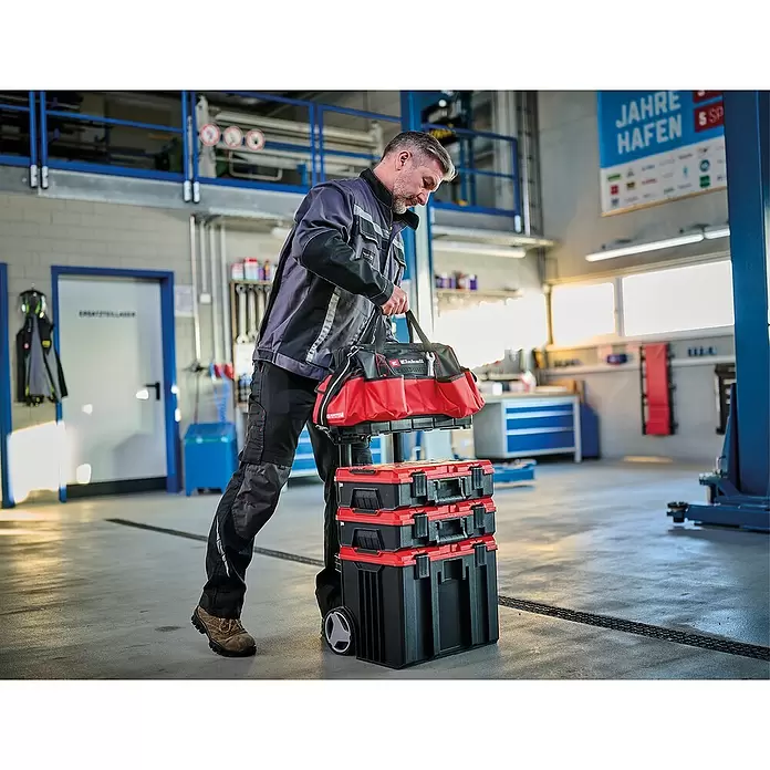 Einhell Werkzeugtasche E-Case - L x B x H: 26,4 x 13,5 x 49 cm, Traglast: 30 kg - Mann trägt eine rot-schwarze Werkzeugtasche, gestapelt auf drei passenden mobilen Werkzeugkisten, in einer Werkstatt.