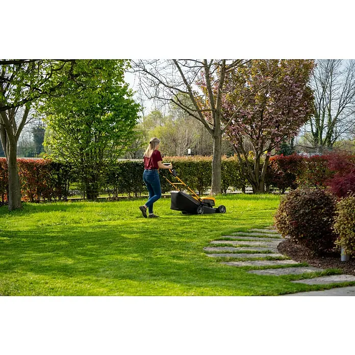 Stiga Akku-Rasenmäher Collector 48 AE Kit - 48 V, 1 Akku, 4 Ah, Schnittbreite: 46 cm - Eine Frau mäht den Rasen mit einem gelben STIGA Rasenmäher in einem grünen Garten mit Bäumen und Sträuchern.