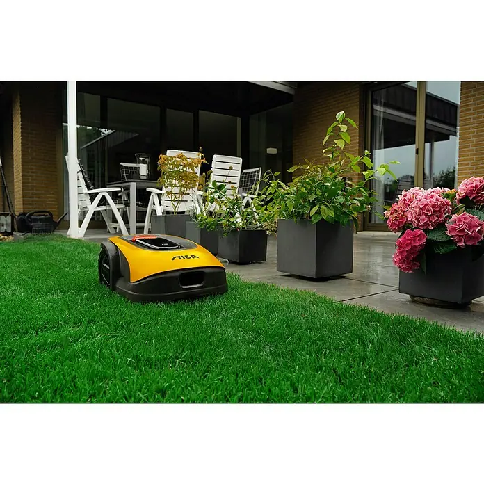 Stiga Mähroboter A750 - 900 m², Schnittbreite: 18 cm, Satellitengestützte Navigation, RTK - Ein gelber Stiga Mähroboter auf einem grünen Rasen vor einer Terrasse mit Pflanzenkübeln.