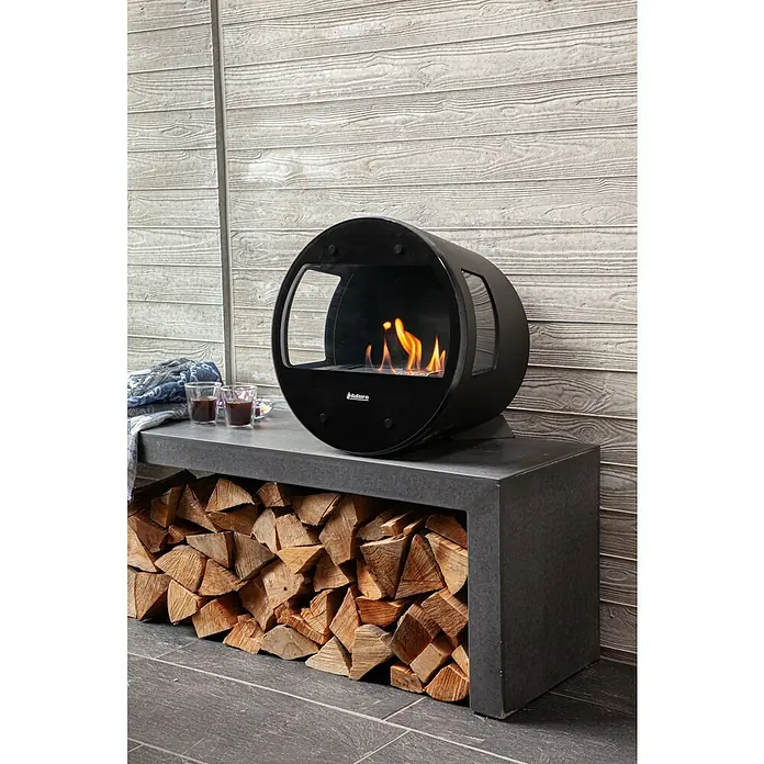Aduro Standfuß - Passend für: Aduro Bioethanol Ofen B1 und B2, Schwarz - Ein moderner, runder, schwarzer Adura-Kaminofen auf einem Regal mit Holzscheiten und zwei Gläsern.