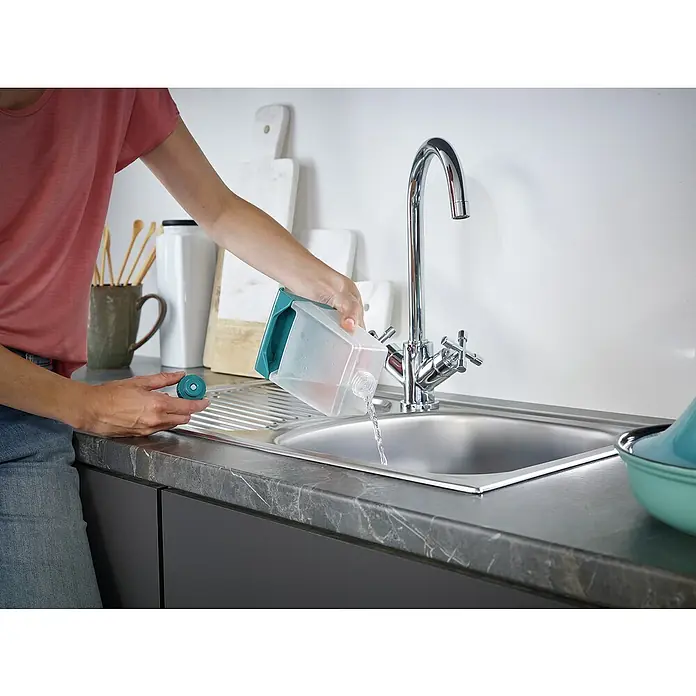 Leifheit Dampfreiniger CleanTenso Power - Aufheizzeit: 30 s, Behältervolumen: 0,68 l - Eine Person leert einen transparenten Wasserfilterkrug mit türkisfarbenem Griff in ein Edelstahlspülbecken.