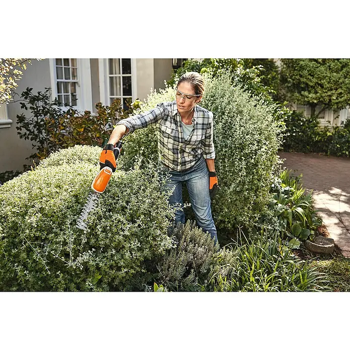 Stihl AS Akku-Strauchschere HSA 26 - 10,8 V, Li-Ionen, 1 Akku - Eine Frau trimmt mit einer orangefarbenen Heckenschere eine Hecke, gekleidet in Jeans und Hemd, mit Schutzbrille und Handschuhen.