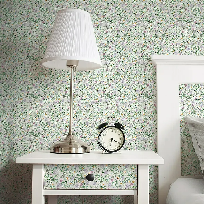Lámina decorativa autoadhesiva vinílico Premium - 90 x 280 cm, Lulu, Verde - Primer plano de una lámpara de noche y un despertador en una mesita de noche con cajón frente a un papel pintado floral.
