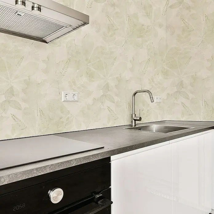 Papel pintado de vinilo autoadhesivo Demeter - Beige, L x An: 280 x 65 cm - Vista de la cocina con fregadero, encimera y fogones bajo una campana extractora frente a un papel pintado con motivos de hojas.