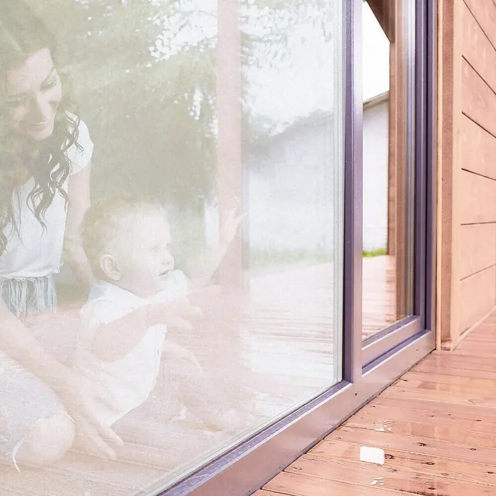 Lámina para ventana de vinilo Cosmos - 67,5 cm x 2 m, Transparente - Primer plano de una puerta de patio moderna con marco oscuro, que ofrece vistas al exterior y un reflejo de una madre con un niño.