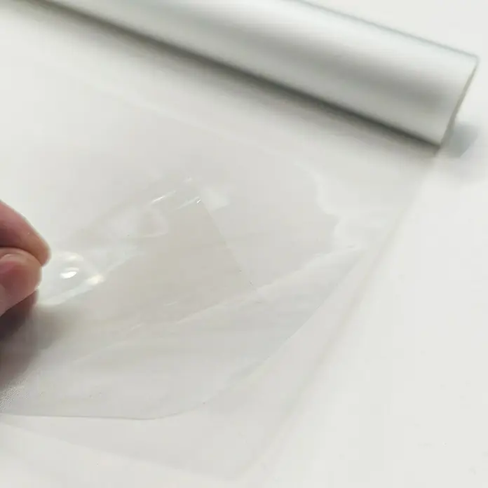 Lámina para ventana de vinilo Cosmos - 67,5 cm x 2 m, Transparente - Primer plano de un rollo de film adhesivo transparente, parcialmente desenrollado. Ideal para embalaje, protección y manualidades.