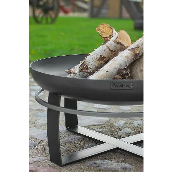 CookKing Feuerschale Viking - Durchmesser: 60 cm, Stahl - Schwarze Cook King Feuerstelle aus Metall mit Birkenholzscheiten, draußen auf Pflastersteinen.