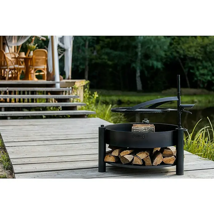 CookKing Feuerschale Montana X - Durchmesser: 88 cm, Stahl - Schwarze Metall-Feuerschale mit Grillaufsatz und Holzfach, auf einer Holzterrasse am Teich.