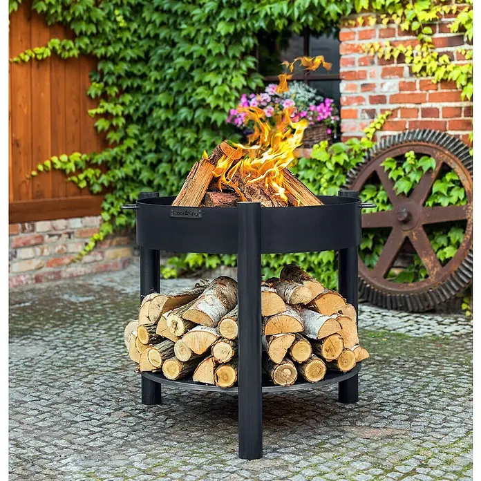 CookKing Feuerschale Montana - Ø x H: 88 x 80 cm, Stahl - Schwarze Feuerschale mit integriertem Holzregal, brennendem Holz, auf Pflastersteinen umgeben von Efeu und Ziegeln.