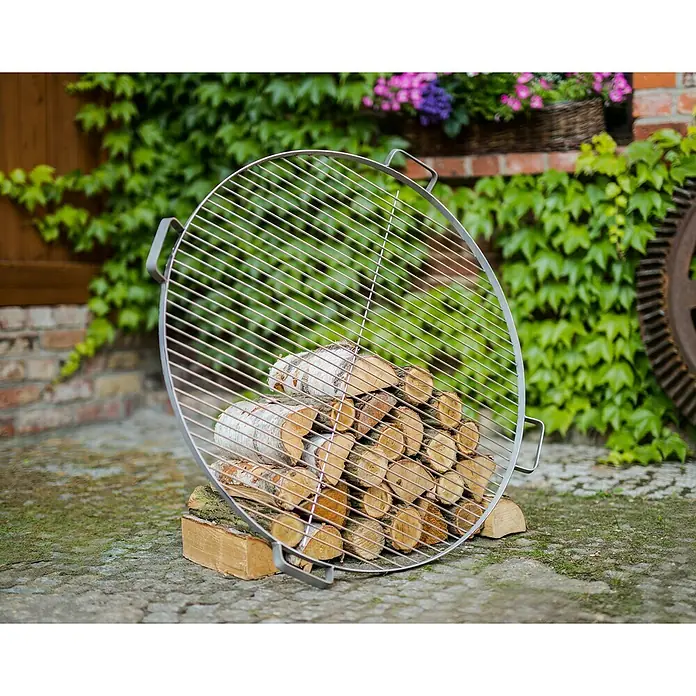 CookKing Grillrost - Durchmesser: 80 cm, Edelstahl, Griffe - Runde Metallgrillrost an gestapelten Brennholzstämmen gelehnt. Ideal für das Kochen im Freien.