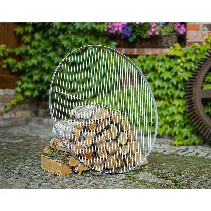 CookKing Grillrost - Durchmesser: 70 cm, Edelstahl - Eine runde, silberne Metall-Grillrostplatte liegt auf einem Holzstapel, was auf eine Outdoor-Kochstelle hindeutet.