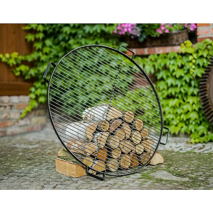 CookKing Grillrost - Durchmesser: 80 cm, Stahl, Griffe - Runde schwarze Metall-Grillrost mit Griffen, lehnt an gestapeltem Brennholz.