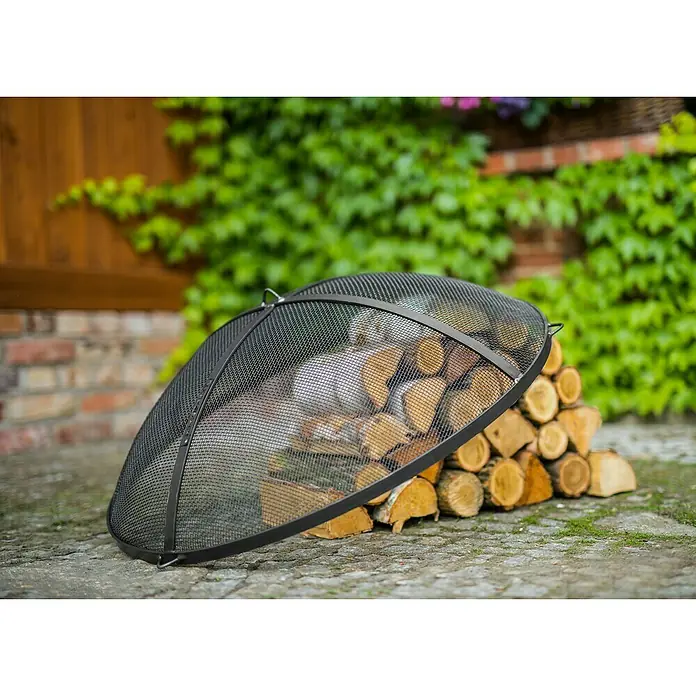 CookKing Funkenschutzgitter - 59 cm, Passend für: Feuerschalen mit Ø 60 cm - Schwarzer Metallgitter-Funkenschutz für Feuerstellen, der im Freien an einem Holzstapel lehnt.