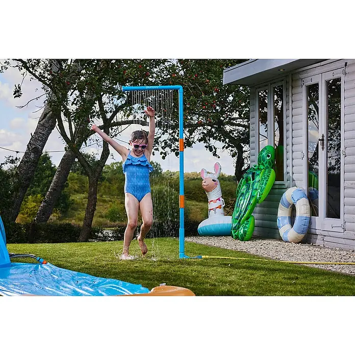 Plum Kinderspielturm Water Park Wasservorhang - Blau - Ein glückliches Kind spielt unter einer blau-orangen Gartendusche/Sprinkleranlage neben einer Wasserrutsche.
