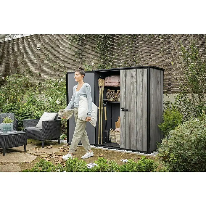 Keter Geräteschrank Signature Strom Grey High-Store - Außenmaß inkl. Dachüberstand (B x T): 140 x 73,6 cm, Stormgrey, Kunststoff - Graue Outdoor-Gartenschrank in Holzoptik im Garten geöffnet, mit Kissen, Fackeln und Gartengeräten. Eine Frau geht vorbei.