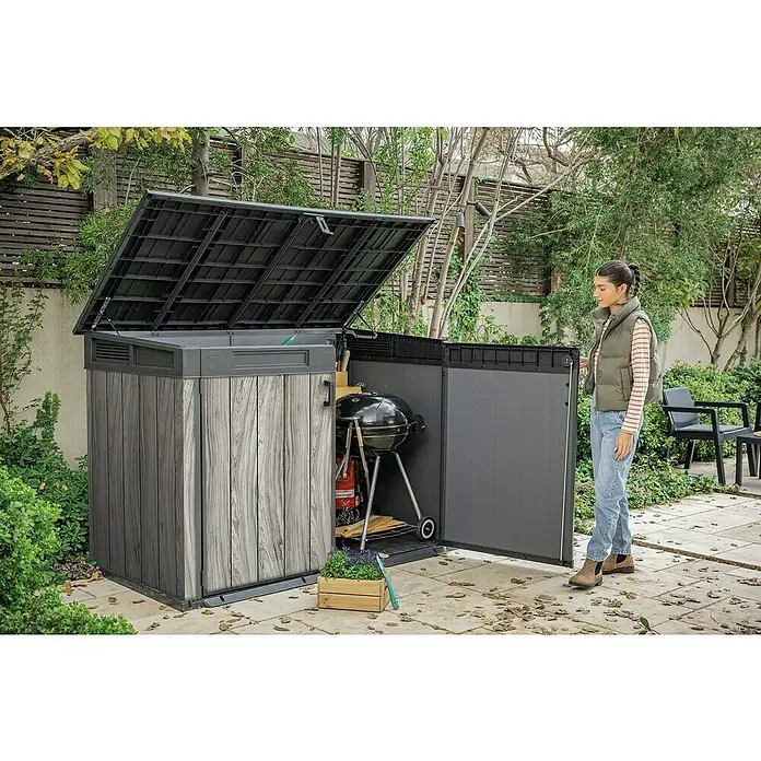 Keter Garten-Aufbewahrungsbox Signature Storm Grey Grande-Store - Außenmaß inkl. Dachüberstand (B x T): 190 x 109 cm, Kunststoff, Stormgrey - Großer Outdoor-Lagerschuppen mit dunklen Holzmaserungsseiten und schwarzem Deckel, offen für Grill im Garten, eine Frau öffnet ihn.
