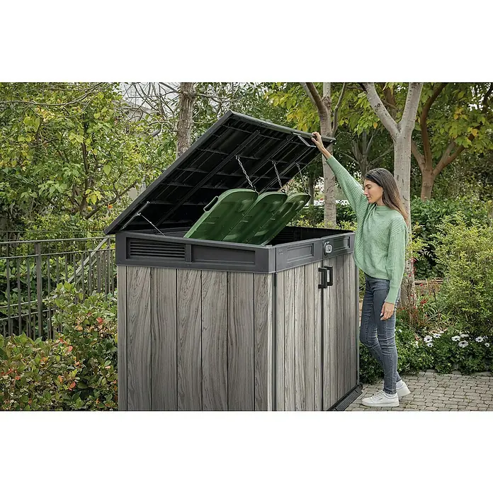 Keter Garten-Aufbewahrungsbox Signature Storm Grey Grande-Store - Außenmaß inkl. Dachüberstand (B x T): 190 x 109 cm, Kunststoff, Stormgrey - Frau öffnet einen grauen Outdoor-Stauraum in Holzoptik mit schwarzem Deckel, der zwei grüne Mülltonnen im Garten zeigt.