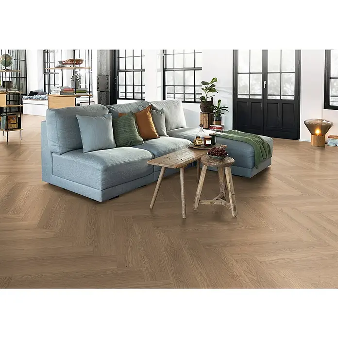 Egger Laminat NatureSense Herringbone - Baronia Eiche braun, Fischgrät, 840 x 168 x 8 mm - Modernes Wohnzimmer mit einem hellen Sofa, Holzboden im Fischgrätmuster und einem rustikalen Holztisch. Die Einrichtung strahlt Gemütlichkeit aus.