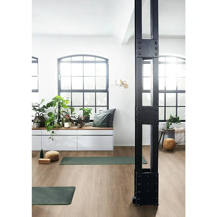 Egger Laminat NatureSense - Newport Eiche braun, Landhausdiele, 1.292 x 193 x 8 mm - Eine helle, moderne Yoga-Studio-Szene mit viel Tageslicht durch große Fenster und einer schwarzen Metallsäule im Vordergrund.
