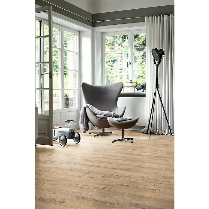 Egger Laminat NatureSense - Sonnenberg Fichte vintage, Landhausdiele, 1.292 x 193 x 8 mm - Einladendes Interieur mit einem grauen Sessel und Hocker auf einem warmen Holzboden, beleuchtet von einer eleganten Stehlampe neben transparenten Vorhängen.