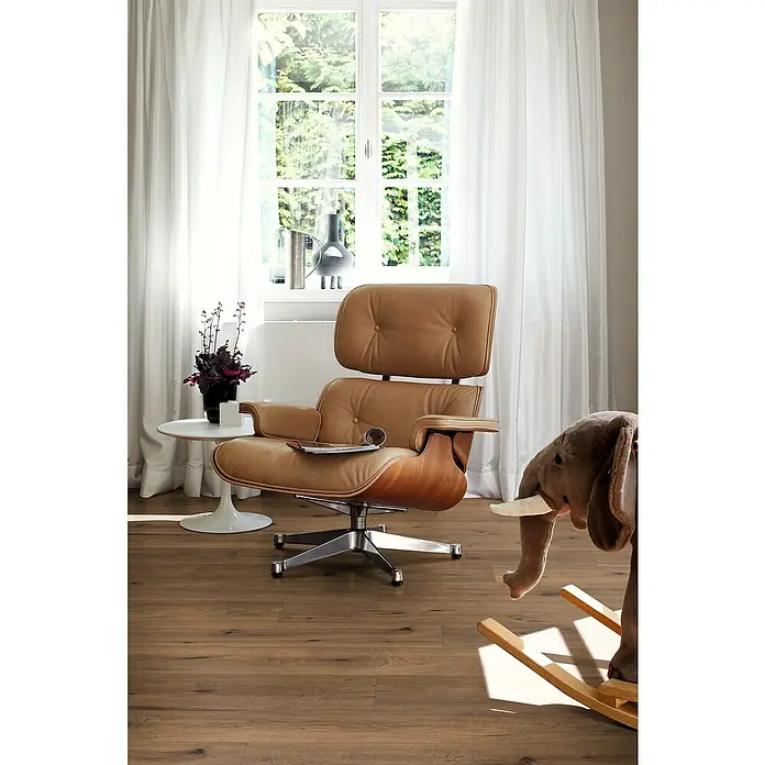 Egger Laminat NatureSense - Inuvik Eiche, Landhausdiele, 1.292 x 193 x 7 mm - Luxuriöser Sessel aus braunem Leder mit passendem Beistelltisch und Spielzeug-Elefant vor einem hellen Fenster.