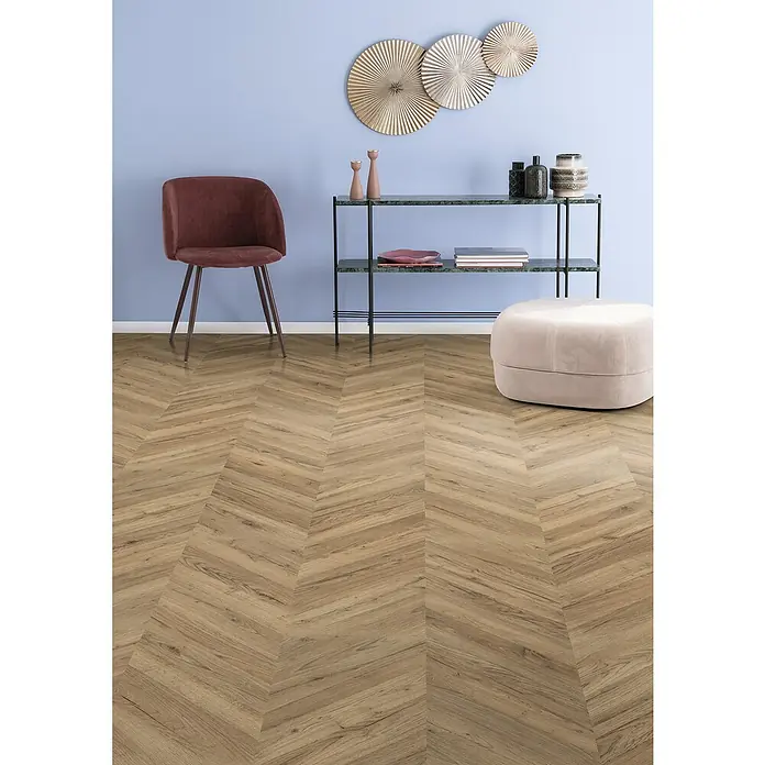 Egger Laminat NatureSense - Rillington Eiche dunkel, Fischgrät, 1.292 x 327 x 8 mm - Ein Interieur mit einem eleganten braunen Stuhl, einem Regal mit Deko-Elementen und einem beigen Hocker auf einem stilvollen Fischgrätboden.