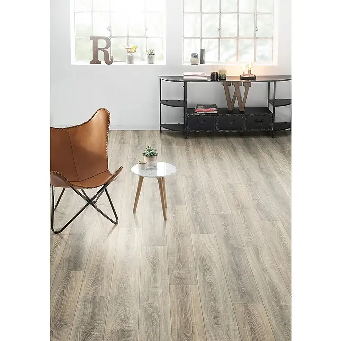Egger Laminat NatureSense - Bardolino Eiche grau, Landhausdiele, 1.292 x 193 x 7 mm - Elegantes Interieur mit hellem Holzboden, einem braunen Ledersessel und einem runden Marmortisch. Eine stilvolle Kommode rundet das Ambiente ab.