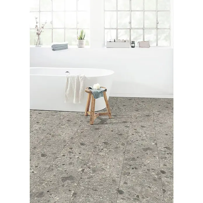 Egger Laminat NatureSense Aqua - Triestino Terrazzo grau, 1.292 x 327 x 8 mm - Badezimmer mit grauen Bodenfliesen im Terrazzo-Stil, Badewanne und Holzhocker. Die Fliesen verleihen dem Raum eine moderne und stilvolle Note.
