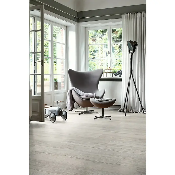Egger Laminat NatureSense Aqua - Elva Eiche grau, Landhausdiele, 1.292 x 193 x 10 mm - Ein heller Raum mit einem grauen Sessel, einer Stehlampe und einem Spielzeugauto auf einem hellen Holzboden.