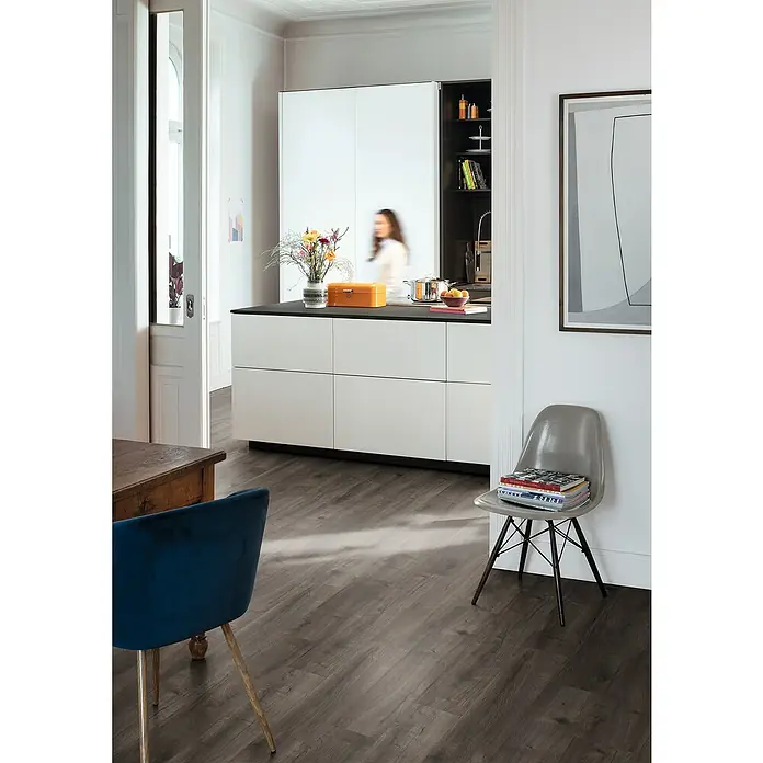 Egger Laminat NatureSense Aqua - Loja Eiche grau, Landhausdiele, 1.292 x 193 x 8 mm - Einblick in eine moderne Küche mit dunklem Holzboden, weißen Schränken und einem grauen Stuhl mit Büchern darauf.