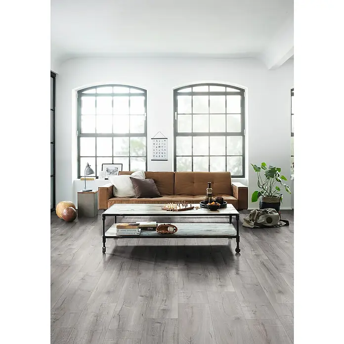 Egger Laminat NatureSense Aqua - Loja Eiche hellgrau, Landhausdiele, 1.292 x 246 x 8 mm - Ein helles Wohnzimmer mit einem robusten Holztisch, der Bücher und Dekorationen trägt, vor einem bequemen Ledersofa.