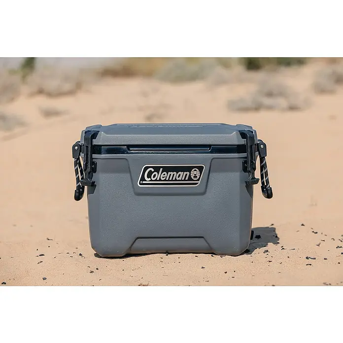 Coleman Kühlbox Convoy QT55 - L x B x H: 37 x 65 x 44 cm, Schwarz, 53 l - Nahaufnahme einer grauen Coleman Kühlbox auf sandigem Untergrund. Die Box ist robust und mit dem Coleman-Logo versehen.