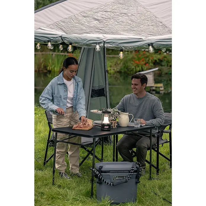 Coleman Kühlbox Convoy QT28 - L x B x H: 26 x 47 x 43 cm, Schwarz, 26,5 l - Eine Coleman Kühlbox steht unter einem Campingtisch, an dem eine Frau Essen zubereitet und ein Mann sitzt.