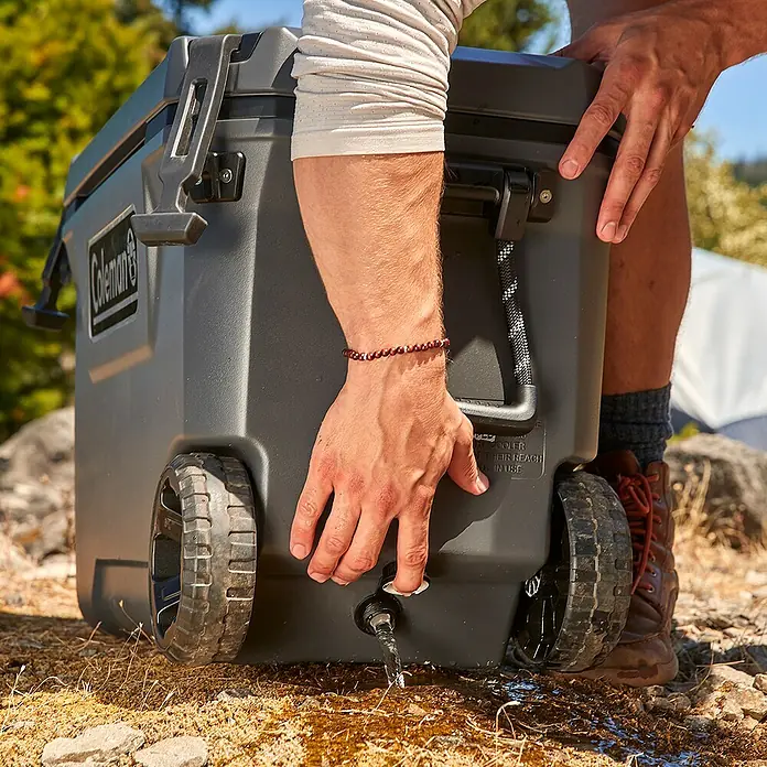 Coleman Nevera de camping Convoy - L x An x Al: 38 x 73 x 47 cm, Negro, 66 l - Primer plano de un refrigerador Coleman con ruedas en el exterior con una persona sacando agua del grifo. Ideal para acampadas.
