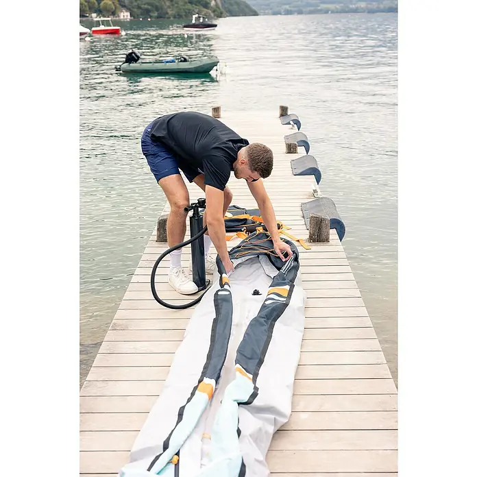 Sevylor Kajak Colorado Kit - L x B: 342 x 96 cm, Nutzlast: 180 kg, Geeignet für: 2 Personen - Ein Mann auf einem Holzsteg pumpt ein aufblasbares Kajak mit einer Handpumpe auf, Boote und ein See sind im Hintergrund.