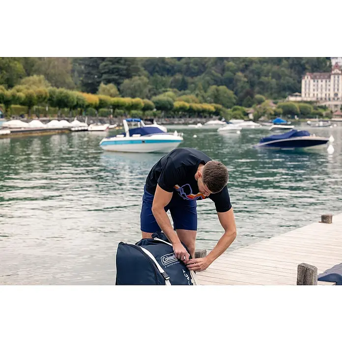 Sevylor Kajak Colorado Kit - L x B: 342 x 96 cm, Nutzlast: 180 kg, Geeignet für: 2 Personen - Ein Mann packt eine blaue Coleman Reisetasche auf einem Holzsteg am See. Im Hintergrund liegen mehrere Boote vor Anker.