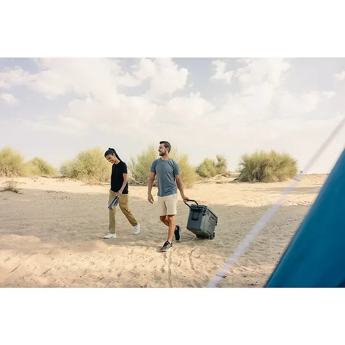 Coleman Kühlbox Convoy QT65 - L x B x H: 38 x 73 x 47 cm, Schwarz, 66 l - Zwei Personen ziehen einen grauen Coleman-Kühlwagen durch eine sandige Landschaft, mit Büschen im Hintergrund und einem Zelt im Vordergrund.